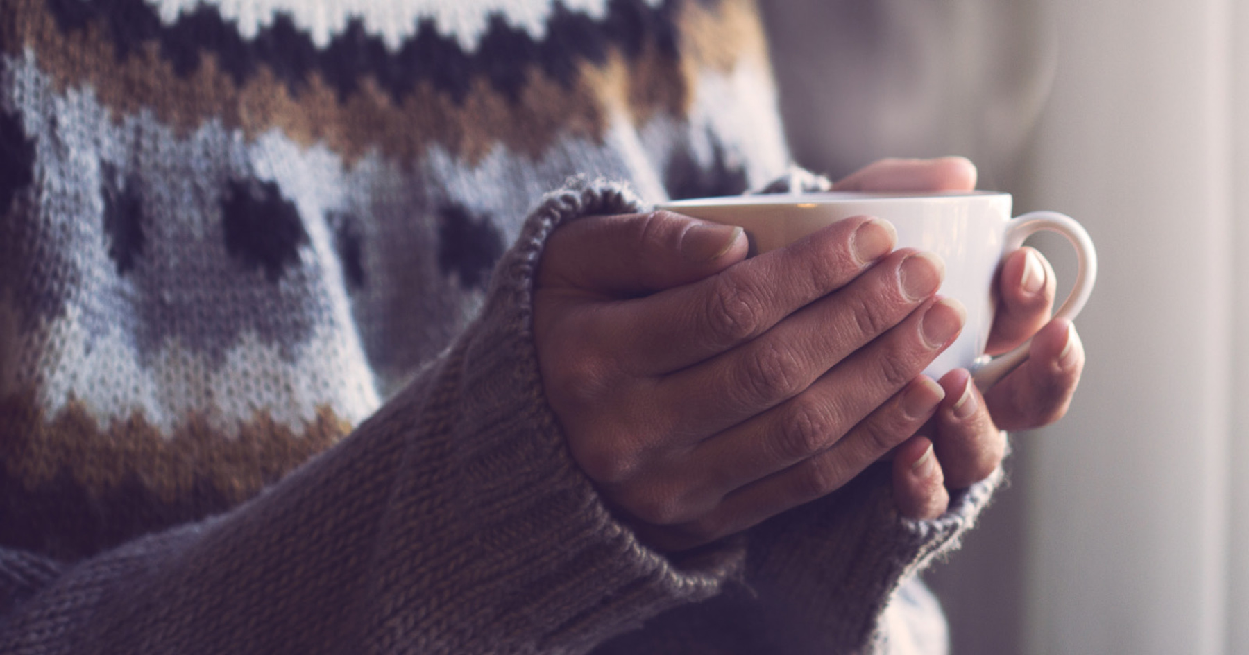 A person in a cozy knitted sweater is holding a white cup with both hands