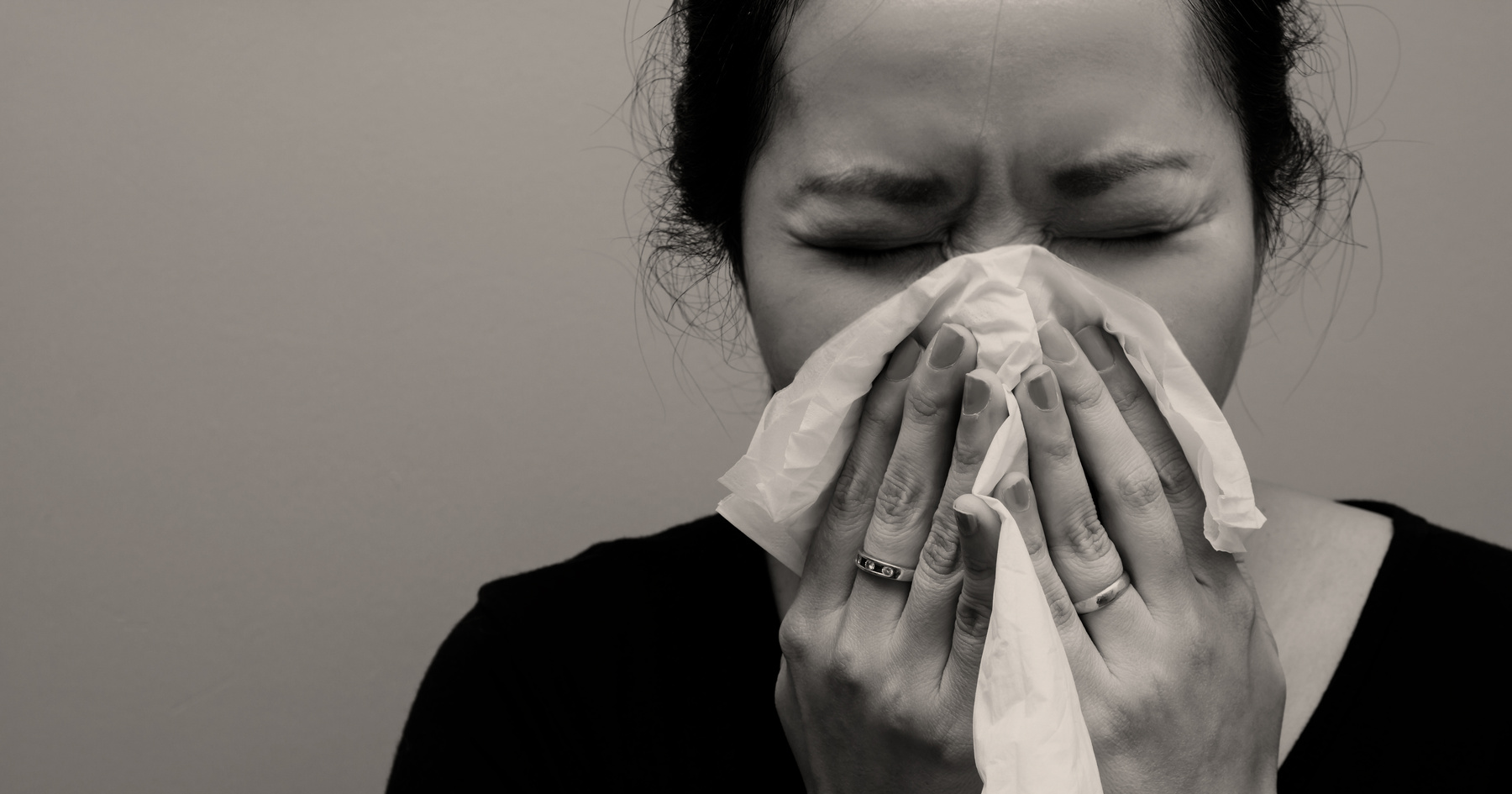 A woman with closed eyes is holding a tissue over her nose and mouth, appearing to sneeze or blow her nose