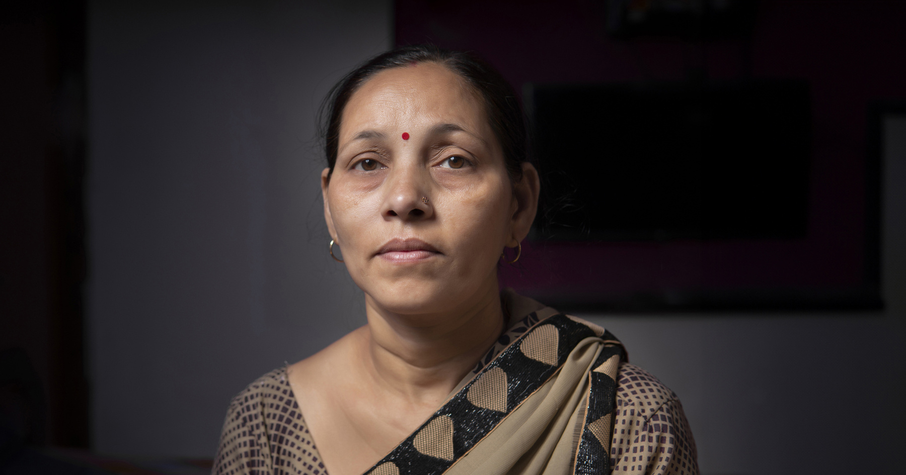 A woman in traditional attire with a calm expression looks directly at the camera in a dimly lit room