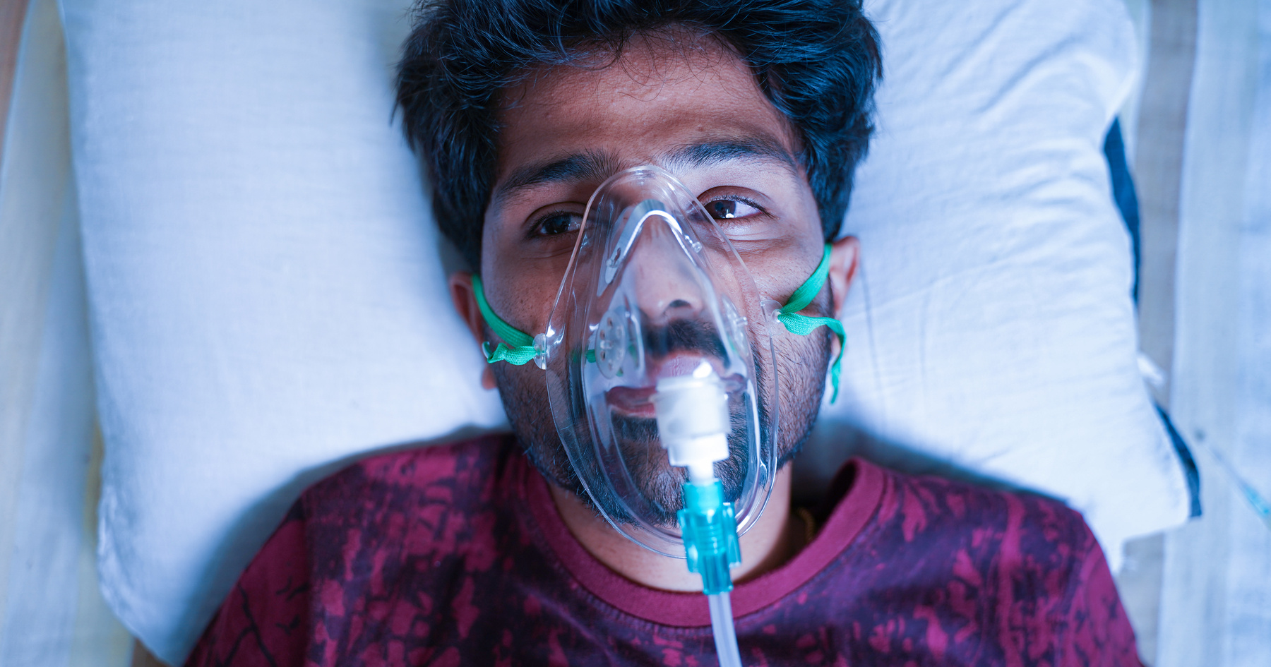 A man lying on a hospital bed wears an oxygen mask, looking upwards