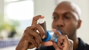 A man wearing a black polo shirt is holding a brown inhaler with a spacer. His hands and inhaler are close up and his face and the background are out of focus.
