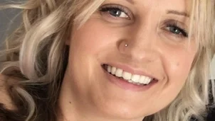 A woman with shoulder length blonde wavy hair is looking directly at the camera and smiling