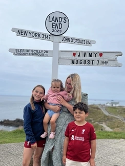 A family of four poses by the Land's End signpost with a coastal view in the background