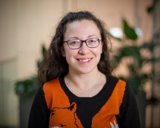 A smiling woman with curly dark hair and glasses wearing a black and orange sweater, with blurred green plants in the background