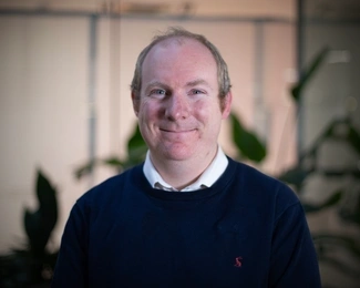 A smiling man with short light brown hair wearing a navy jumper over a white shirt, with blurred green plants in the background