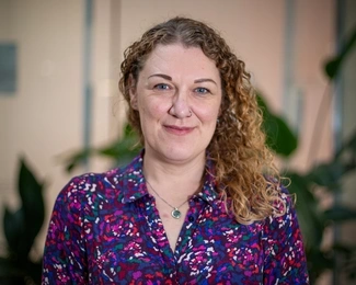 A smiling woman with curly blonde hair wearing a colourful patterned blouse, with blurred green plants in the background