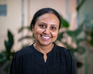 A smiling woman with dark hair pulled back, wearing a black blouse, with blurred green plants in the background