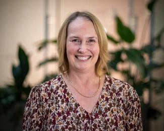 A smiling woman with shoulder-length blonde hair wearing a floral blouse, with blurred green plants in the background