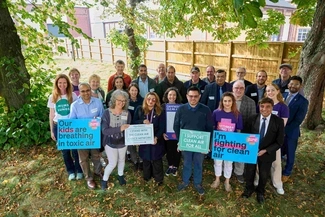 A group of people gathered outdoors holding signs advocating for clean air, with messages supporting the Clean Air Justice Network and Asthma + Lung UK