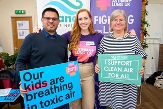 Three people stand together holding signs advocating for clean air, with banners for Asthma + Lung UK and the Clean Air Justice Network in the background