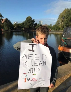 A young boy stands near a canal holding a handmade sign that reads I NEED CLEAN AIR with drawings of cars, trees, and pollution, looking serious