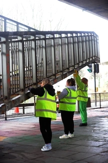 Three people in high-visibility vests clean a metal pedestrian bridge under a motorway