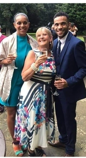 Three smiling people dressed in formal attire hold glasses of champagne at an outdoor event, with a green garden in the background