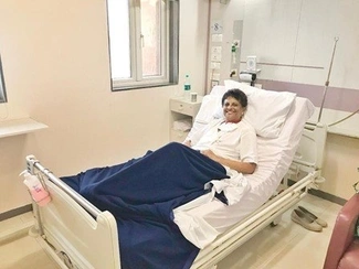 Woman in a hospital bed wearing a white top, smiling, covered with a navy blue blanket in a well-lit hospital room