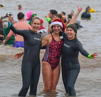 Three smiling women stand in the sea during a festive swim, with two wearing wetsuits and one in a pink swimsuit and Santa hat, surrounded by other swimmers in costume